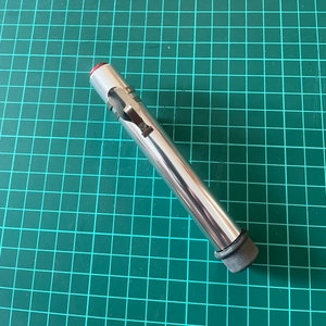 Puede incluir: Un tubo de metal plateado con una tapa roja y una tapa de goma negra. El tubo tiene un pequeño clip de metal en el lateral.