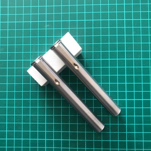 Puede incluir: Dos bolígrafos de metal plateado sobre una estera de corte verde. Los bolígrafos están sobre una superficie blanca.