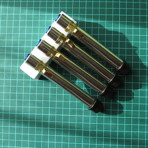 May include: Four silver metal tubes with blue and gold accents are arranged in a white plastic holder. The tubes are arranged in a fan shape, with the tips of the tubes pointing towards the right side of the image.