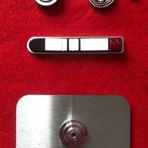 Puede incluir: Una placa de metal plateado con una perilla redonda de plata en el centro. Sobre la placa hay un panel rectangular blanco y negro con una franja roja en el lado derecho. Sobre el panel hay tres piezas de metal plateado: una pieza redonda con un diseño de círculos concéntricos, una pieza redonda con un símbolo en forma de estrella y una pieza pequeña redonda.