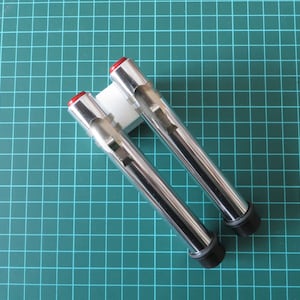 Könnte beinhalten: Zwei verchromte Metallrohre mit roten Kappen und schwarzen Gummikappen. Die Rohre sind auf einem weißen Kunststoffsockel montiert.