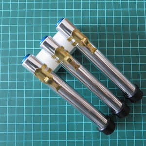 Puede incluir: Tres tubos de metal plateado con puntas de goma negra y detalles de metal dorado. Los tubos están dispuestos en fila y se mantienen unidos por una base de plástico blanco.