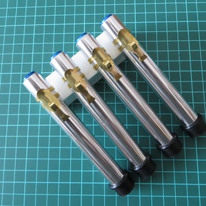 May include: Four silver metal tubes with black caps and gold accents are displayed on a white stand. The tubes are arranged in a row, with the caps facing the viewer.