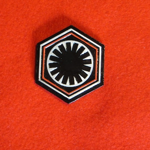 Może przedstawiać: Czarno-biała sześciokątna przypinka z czerwonym obramowaniem. Przypinka przedstawia stylizowany symbol z czarnym kółkiem i białymi promieniami wychodzącymi z centrum.