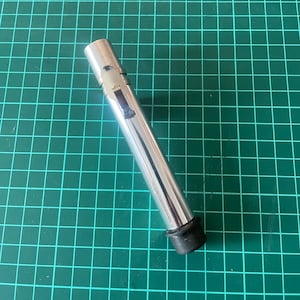 May include: A shiny, cylindrical metal object with a black rubber base. The object has a small rectangular opening and appears to be a tool or part of a larger device. It is lying on a green grid surface.