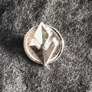 May include: A silver pin featuring a geometric diamond-shaped design. The pin is circular with a raised edge.