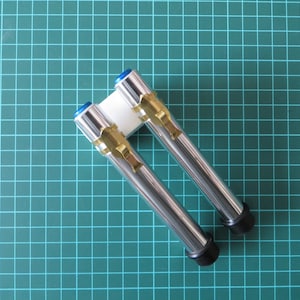 Könnte beinhalten: Zwei silberne und goldfarbene Metallrohre mit blauen Kappen und schwarzen Sockeln. Die Rohre sind an einer weißen Kunststoffbasis befestigt.