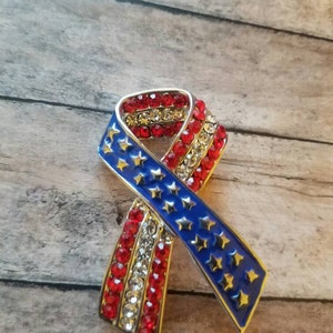 May include: A red, white and blue ribbon pin with rhinestones. The ribbon is in the shape of a bow and has a silver metal frame.