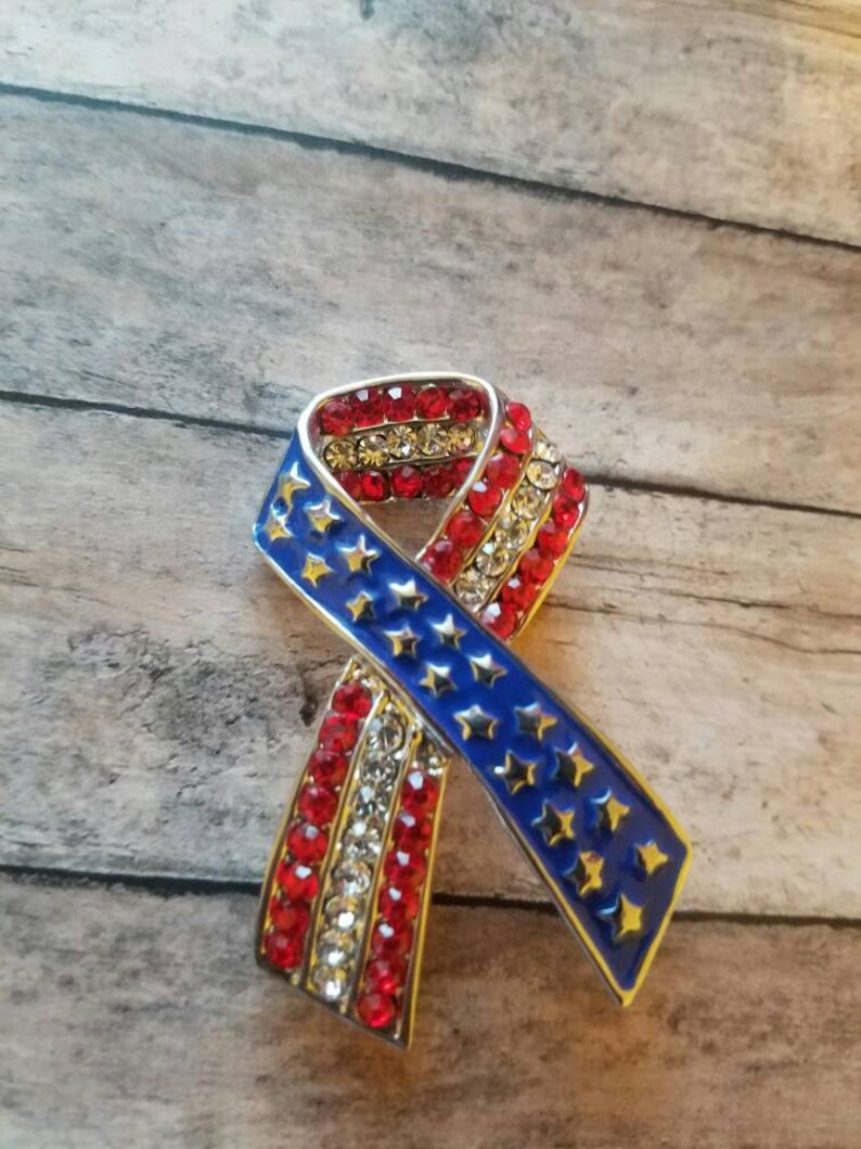 May include: A red, white and blue ribbon pin with rhinestones. The ribbon is in the shape of a bow and has a silver metal frame.