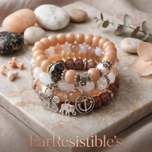 May include: A stack of beaded bracelets featuring peach, white, and brown porous beads. The bracelets include silver-toned charms shaped like a butterfly, an elephant, and an anchor, along with a dark patterned bead and the text EarResistible's.