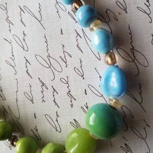 Puede incluir: Un collar hecho de cuentas de vidrio verde y azul con detalles dorados. Las cuentas son redondas y tienen un acabado brillante.
