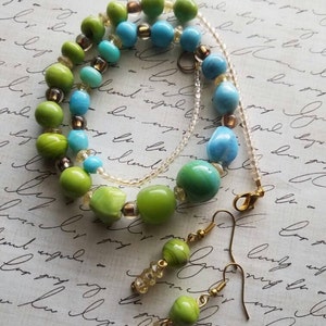 Puede incluir: Un collar y pendientes a juego de cuentas de vidrio verde y azul. El collar tiene una cadena plateada y los pendientes tienen ganchos dorados.