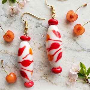 May include: A pair of dangle earrings featuring elongated white glass beads with red and orange abstract patterns. Each bead is accented with small red glass spacers and gold-tone metal hooks. The text EarResistible's is printed at the bottom.