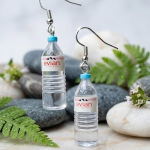 May include: A pair of dangle earrings featuring miniature clear plastic water bottles with light blue caps and white labels printed with the text 'evian' in red and a black mountain graphic, suspended from silver-toned metal earring hooks.