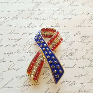 May include: A red, white and blue ribbon pin with rhinestones. The ribbon is in the shape of a bow and has stars on it.