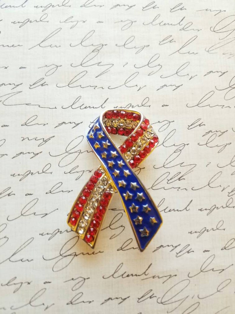 May include: A red, white and blue ribbon pin with rhinestones. The ribbon is in the shape of a bow and has stars on it.