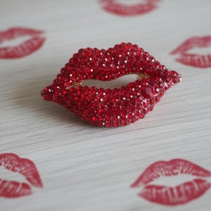 May include: A red rhinestone lips brooch with a gold-tone backing. The brooch is surrounded by red lip print designs and the text "EarResistible's" at the bottom. The brooch is a statement piece.
