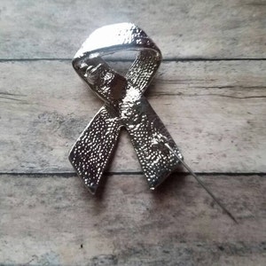 May include: Silver ribbon pin with a textured finish. The pin is in the shape of a ribbon tied in a bow.