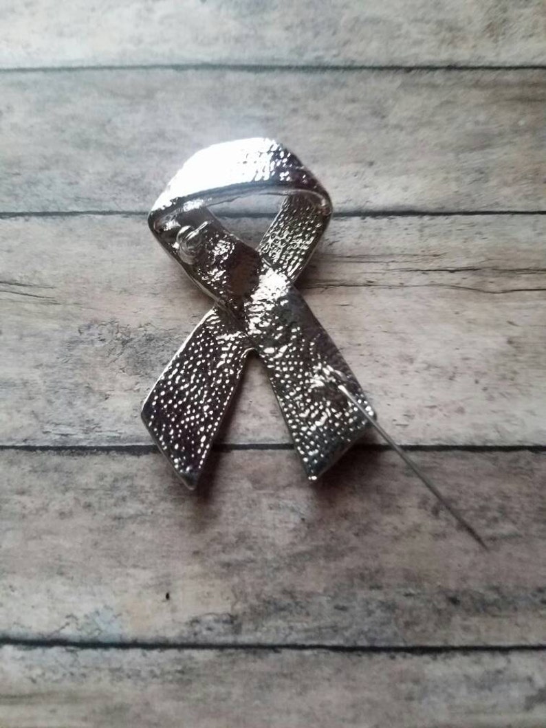 May include: Silver ribbon pin with a textured finish. The pin is in the shape of a ribbon tied in a bow.