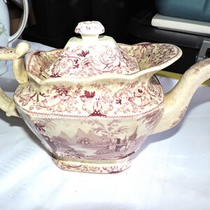 Peut inclure: Une théière vintage de couleur crème avec un motif floral rouge et un corps carré. La théière a un couvercle avec un bouton sur le dessus et un bec verseur sur le côté. La théière est décorée d'un motif floral rouge et d'une scène de paysage avec des bâtiments.