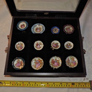 May include: A wooden box with a glass lid containing 12 miniature porcelain plates with floral and romantic designs. The plates are arranged in a grid pattern and are displayed on a black velvet background.