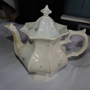 May include: White porcelain teapot with a green and blue floral design. The teapot has a lid with a pointed top and a handle with a decorative scroll design.