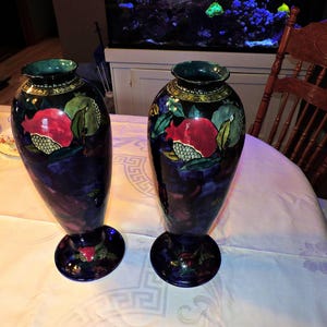 May include: Two dark blue ceramic vases with floral and fruit designs. The vases have a glossy finish and are decorated with red, green, and yellow accents. The vases are sitting on a white tablecloth.