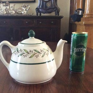 May include: A white ceramic teapot with a green finial and a green and gold leaf pattern. A green can of Perrier sits beside the teapot on a wooden table. The teapot features a curved handle and a spout.