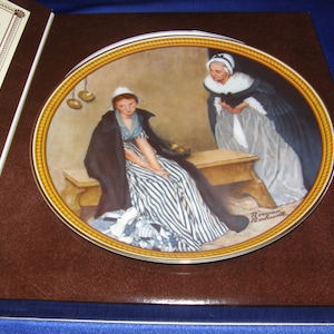 May include: A porcelain plate depicting a painting of two women in period dress. The woman on the left is wearing a striped dress and a black shawl. The woman on the right is wearing a white dress and a black shawl. The plate has a gold rim and the artist's signature, "Norman Rockwell", is visible in the bottom right corner.