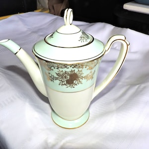 May include: A white and green ceramic coffee pot with gold trim and floral designs. The pot has a lid with a knob on top.