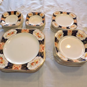 May include: A set of square-shaped ceramic plates with a blue, gold, and orange floral pattern. The plates are stacked in groups of four.