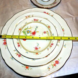 May include: Set of three white ceramic dinner plates with a floral pattern in shades of pink, yellow, blue, and green. The plates have a gold trim and are nested together.