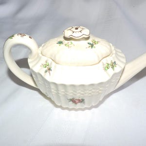 May include: A white ceramic teapot with a floral design and a lid with a floral knob. The teapot has a fluted design and a spout on the side.