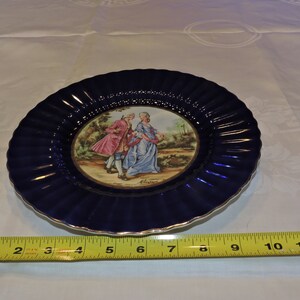 May include: A blue and white porcelain plate with a gold rim. The plate features a central image of a couple in 18th-century clothing walking in a garden. The plate has a scalloped edge.