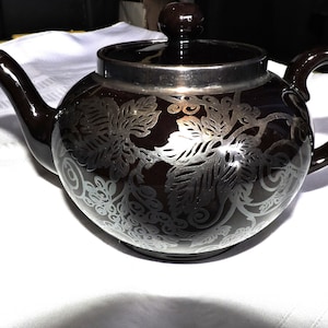 Peut inclure: Une théière brune avec un motif floral argenté. La théière a un couvercle et une poignée.