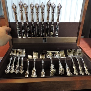 May include: A wooden box with a velvet lining containing a set of silver cutlery. The cutlery is arranged in rows and includes knives, forks, and spoons.