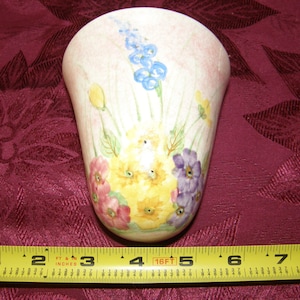 Peut inclure: Un vase mural en céramique blanche avec un motif floral. Le vase présente un motif de fleurs roses, jaunes et violettes avec des feuilles vertes. Le vase mesure environ 10 cm de haut.