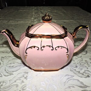 May include: A pink teapot with gold trim and a decorative design. The teapot has a lid with a gold knob and a spout on the side.