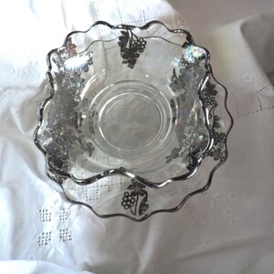 May include: A clear glass bowl with a scalloped edge and silver grapevine design. The bowl is on a white tablecloth.