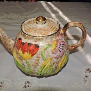 May include: A brown teapot with a floral design featuring red, yellow, and pink flowers. The teapot has a gold lid and a brown handle.