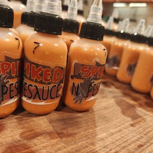 May include: Bottles of orange-colored sauce with black caps and white nozzles. The labels read "Spiked N Pest Sauce" in red and brown lettering, with a saw blade graphic. The number "7" is written on some bottles.