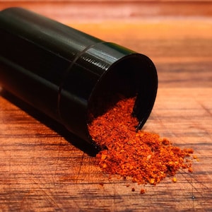 May include: A black plastic container with a red and orange spice blend spilling out onto a wooden surface.