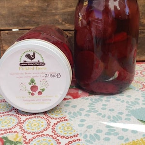 May include: Two glass jars filled with pickled beets. The jars have white lids and are sitting on a floral tablecloth. The label on one jar reads "Farmer Little's This N'at Pickled Beets" and lists the ingredients: beets, water, white sugar, onion, garlic. The lot number is 2110043.