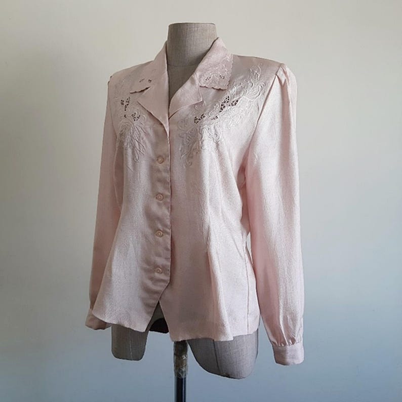 May include: A pale pink long-sleeved blouse with a collared neckline and button closure. The blouse has embroidered floral detailing on the collar and chest. The fabric has a textured look, and the sleeves have buttoned cuffs. The blouse is displayed on a mannequin.