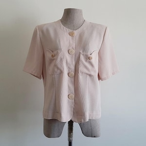 Op de afbeelding: Een lichtroze blouse met korte mouwen, een ronde halslijn, vier grote ronde knopen aan de voorzijde en twee borstzakken met kleinere bijpassende knopen op elke klep, vervaardigd uit een stof met een textuur.