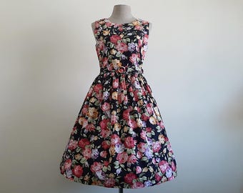 May include: A sleeveless dress featuring a black background with a vibrant floral print of red, pink, yellow, and purple roses with green leaves. The dress has a round neckline, a fitted bodice, and a full, pleated skirt with a matching belt.