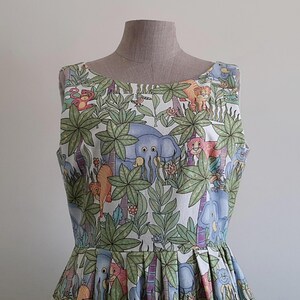 May include: A sleeveless dress featuring a jungle-themed print with elephants, lions, monkeys, and lush green foliage. The dress is displayed on a mannequin, showcasing its fitted bodice and pleated skirt. The fabric has a white background with colorful illustrations.
