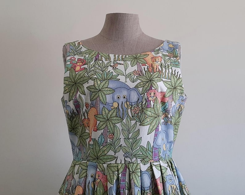 May include: A sleeveless dress featuring a jungle-themed print with elephants, lions, monkeys, and lush green foliage. The dress is displayed on a mannequin, showcasing its fitted bodice and pleated skirt. The fabric has a white background with colorful illustrations.