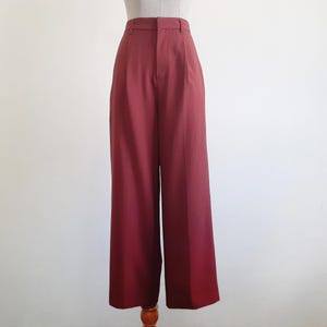 May include: Wide-leg trousers in a deep burgundy hue. The high-waisted design features a button and zip fastening, belt loops, and pleats at the waist. The fabric appears to be a smooth, woven material. The trousers are displayed on a mannequin.