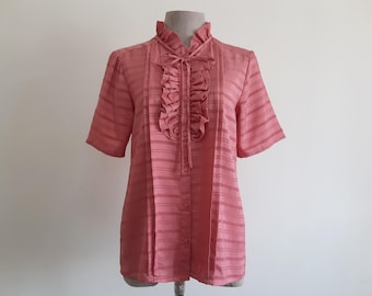 May include: A short-sleeved, salmon-pink button-down blouse featuring a high ruffled collar, a vertical ruffled placket down the center front, and a delicate tie closure at the neckline. The fabric has a subtle horizontal striped texture.
